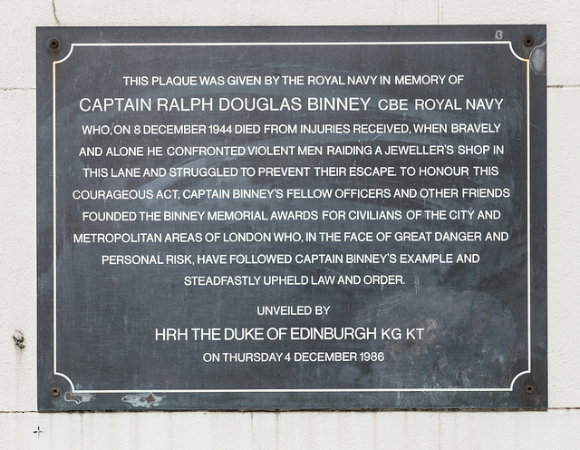 Ralph Douglas Binney black plaque - This plaque was given by the Royal Navy in memory Captain Ralph Douglas Binney CBE Royal Navy who, on 8 December 1944 died from injuries received, when bravely and alone he confronted violent men raiding a jeweller's shop in this lane and struggled to prevent their escape. To honour this courageous act, Captain Binney's fellow officers and other friends founded the Binney Memorial Awards for civilians of the City and Metropolitan areas of London who, in the face of great danger and personal risk