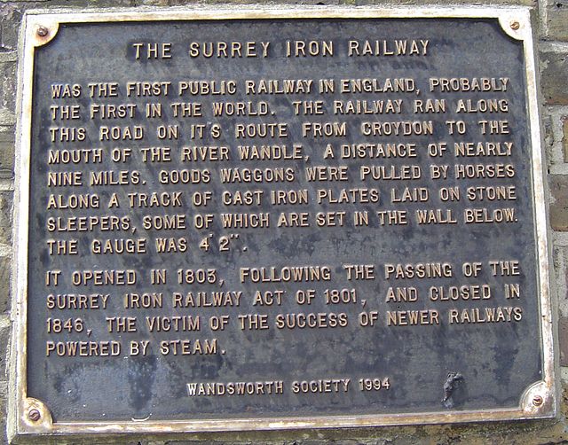 Surrey Iron Railway brass plaque - The Surrey Iron Railway was the first public railway in England, probably in the world. The railway ran along this road on its route from Croydon to the mouth of the River Wandle, a distance of nearly nine miles. Goods wagons were pulled by horses along a track of cast iron plates laid on stone sleepers, some of which are set in the wall below. The gauge was 4'2. It opened in 1803, following the passing of the Surrey Iron Railway Act in 1801, and closed in 1846, the victim of the success of newe